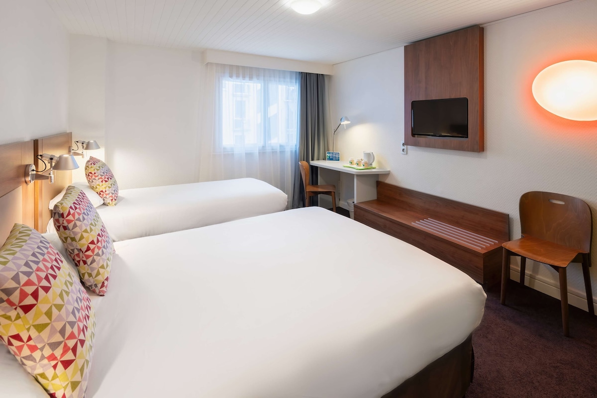 Une chambre d’hôtel avec deux lits, un bureau, une chaise, une télévision et une fenêtre avec des rideaux.