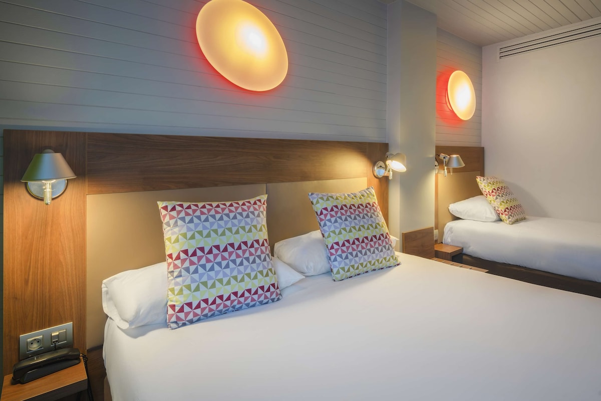 Une chambre d’hôtel avec deux lits, chacun doté d’une tête de lit en bois, de draps blancs et d’oreillers aux motifs géométriques colorés. On y trouve des lampes fixées au mur et un téléphone sur la table de chevet.