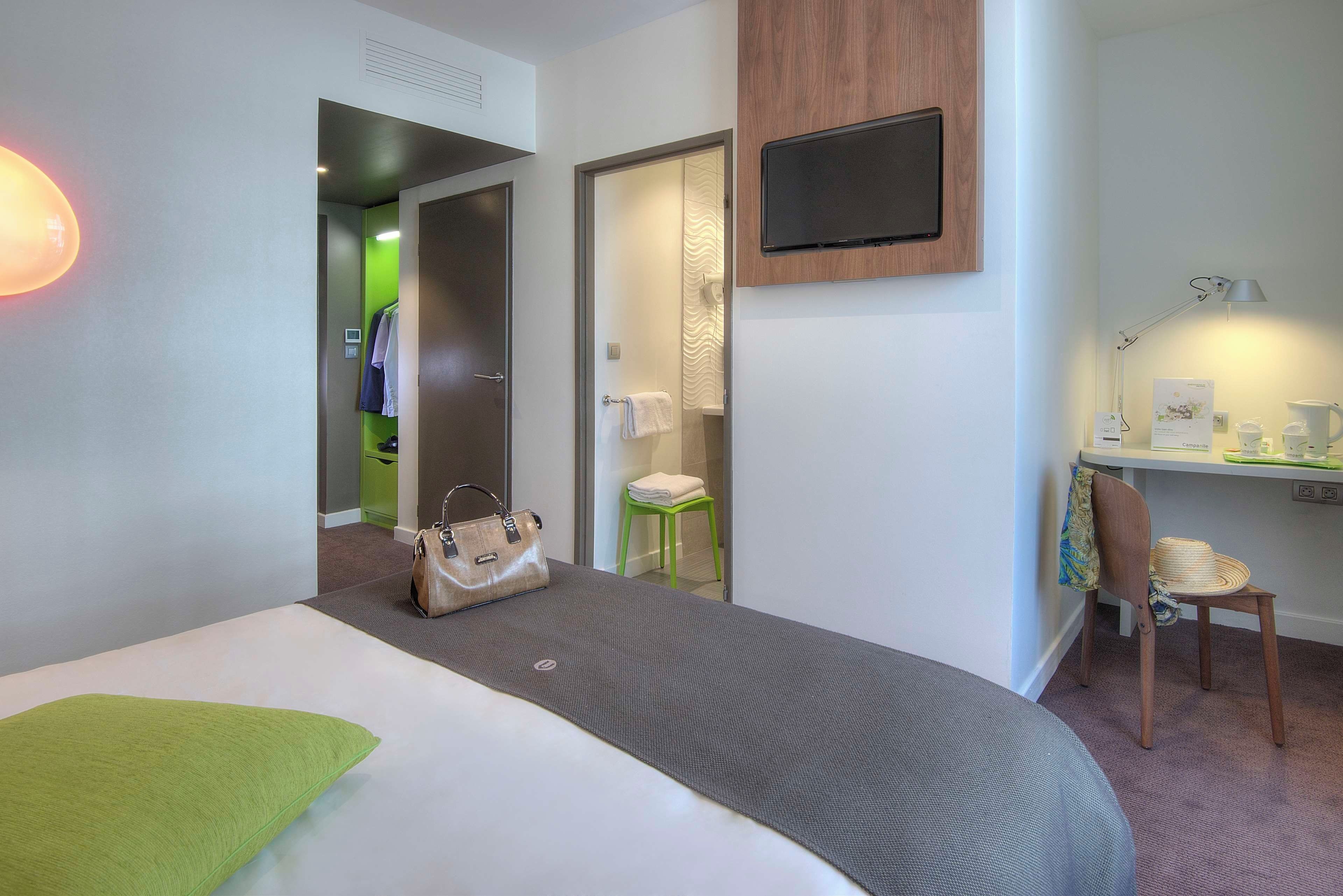 A hotel room with a bed, a desk with a chair, a TV mounted on the wall, and a bathroom visible through an open door.