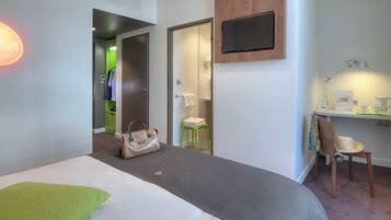 A hotel room with a bed, a desk with a chair, a TV mounted on the wall, and a bathroom visible through an open door.
