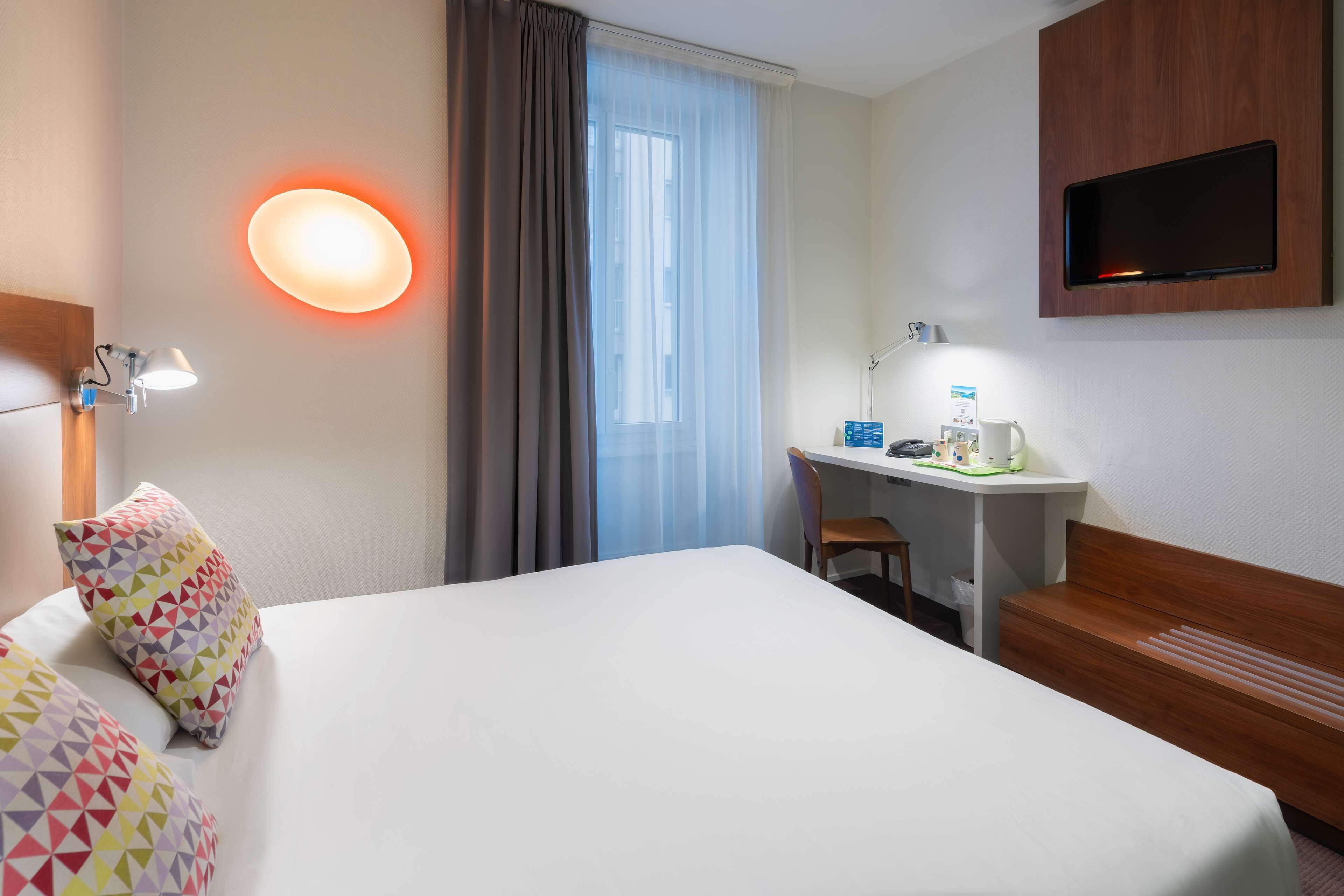 A hotel room with a bed, a desk with a chair, a television, and a window with curtains.