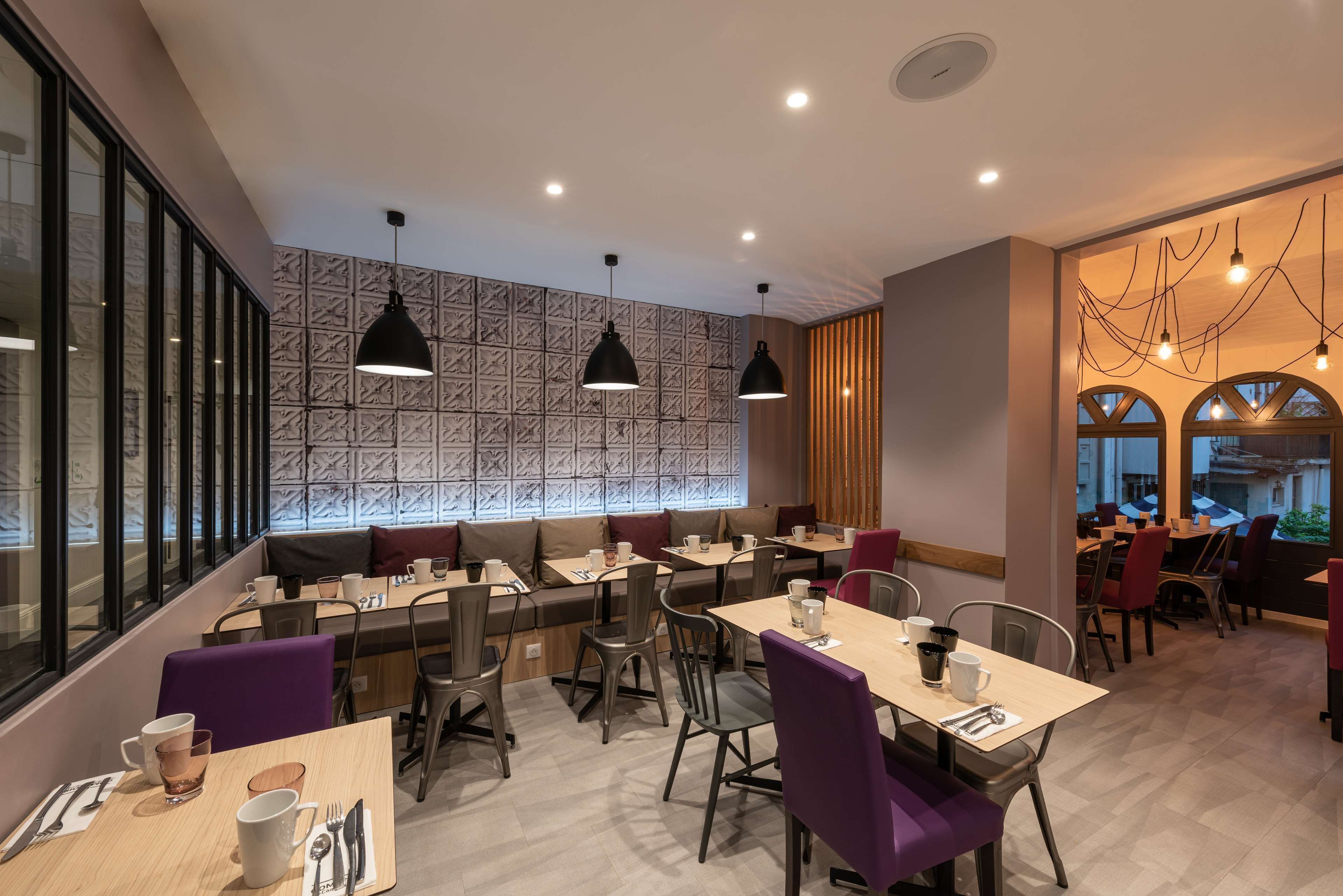 Un café moderno con varias zonas de estar, incluyendo una mesa común alargada y mesas individuales con sillas.