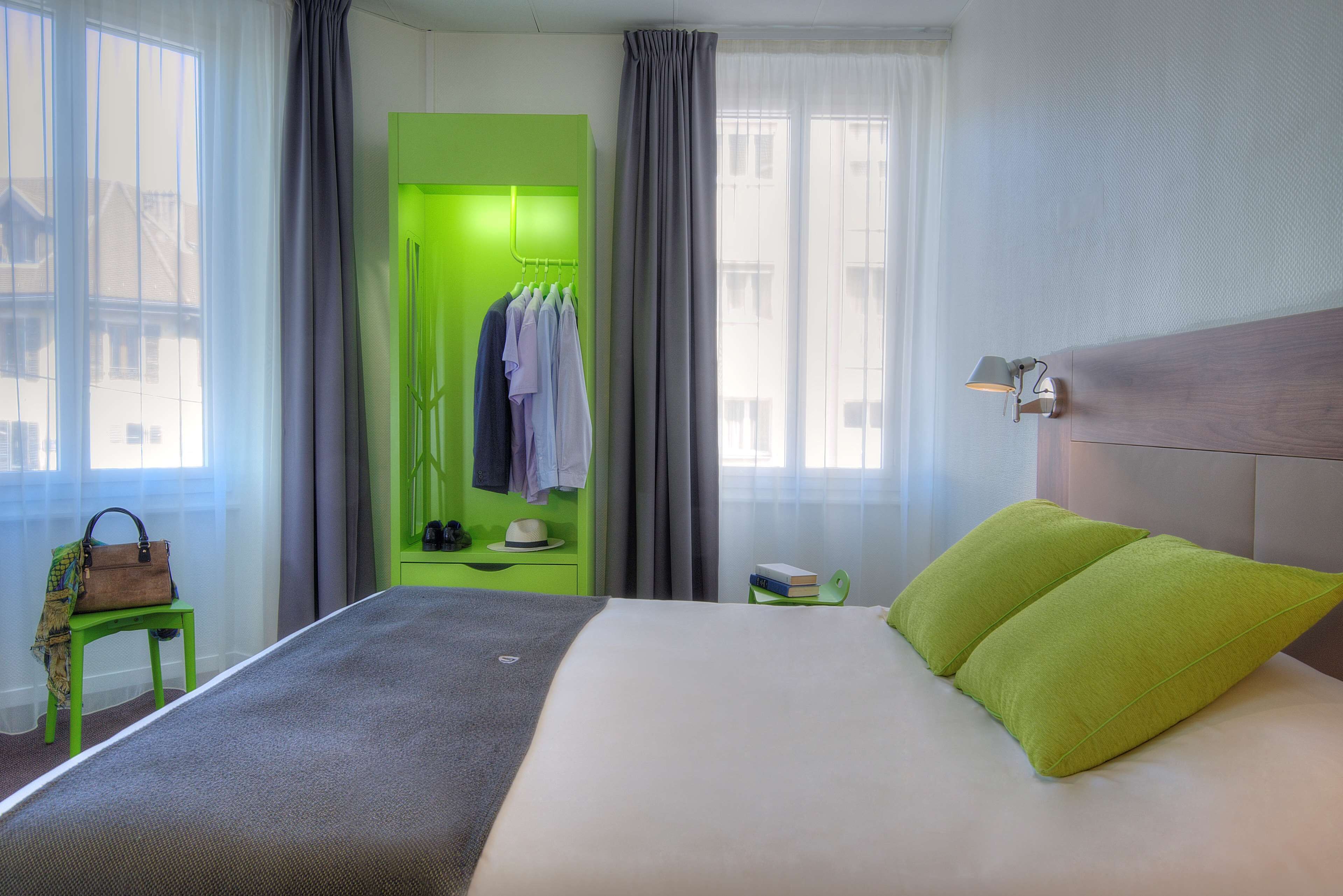 Una camera d'albergo con un letto, un armadio verde, un tavolino con una borsa e un comodino con una lampada.