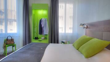 Una habitación de hotel con una cama, un armario verde, una pequeña mesa con un bolso y una mesita de noche con una lámpara.