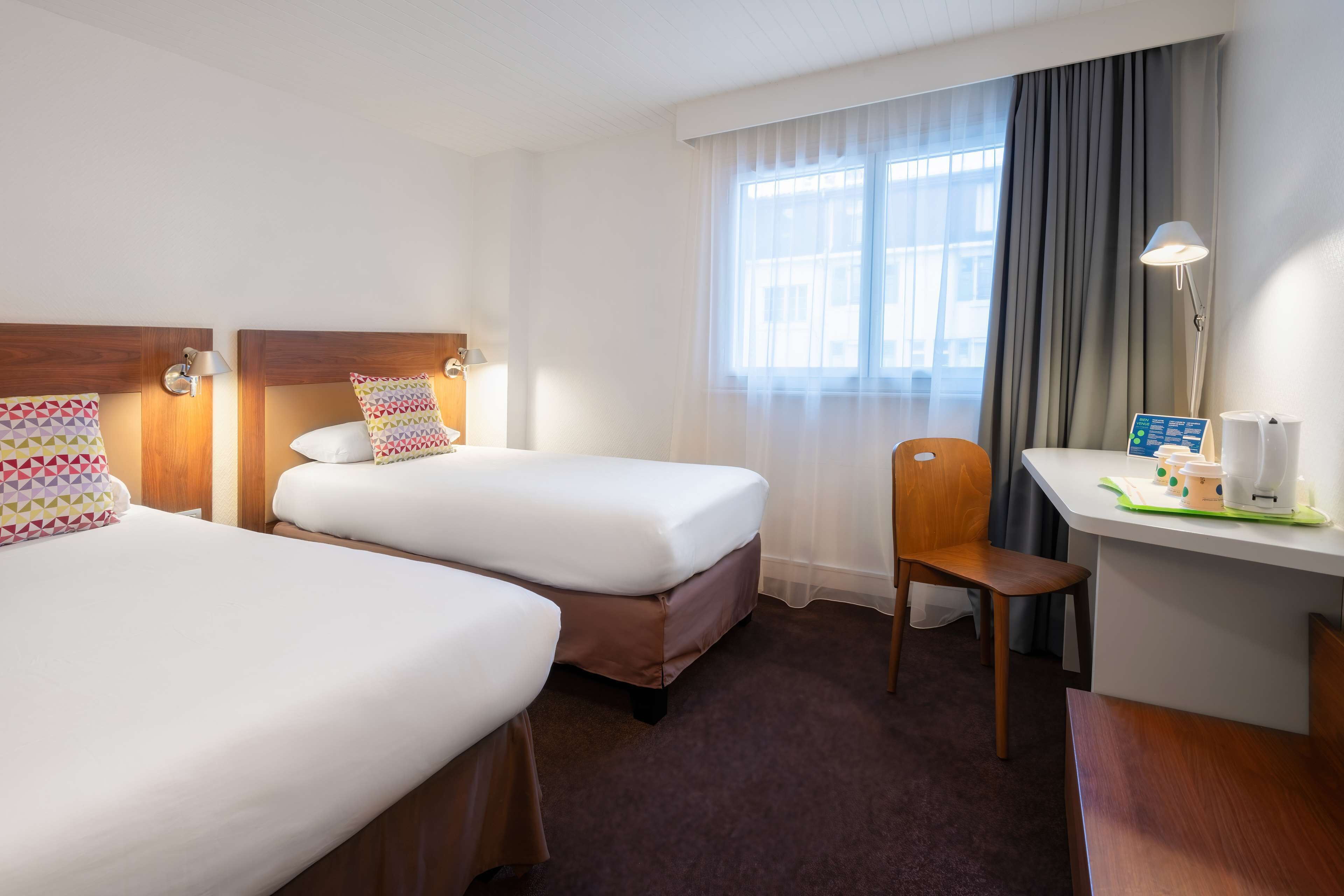 A hotel room with two beds, a desk, a chair, a lamp, and a window with curtains.