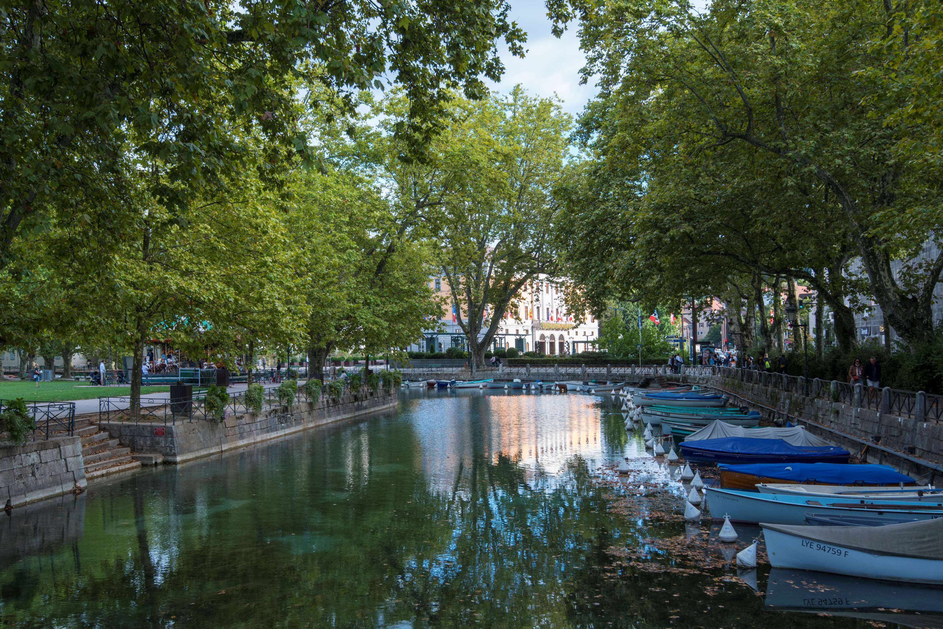 Un río tranquilo con barcas amarradas a la derecha, rodeado de árboles y un muro de piedra.