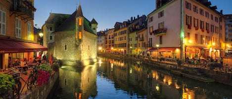 A historic stone building with a tower, reflecting in a calm canal, surrounded by other buildings with outdoor seating and flower arrangements.