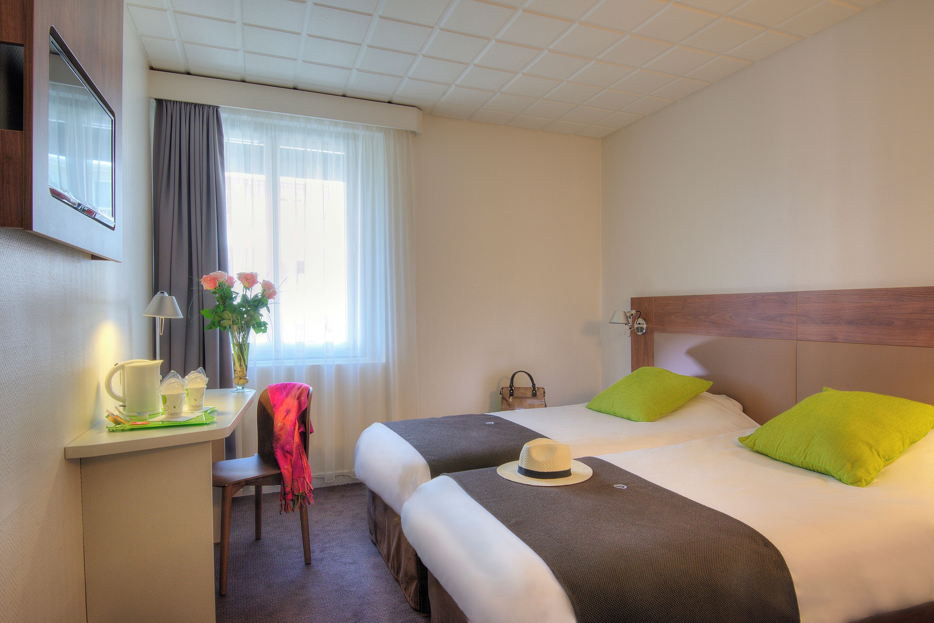 A hotel room with two beds, a desk with a lamp, a chair, and a window with curtains.