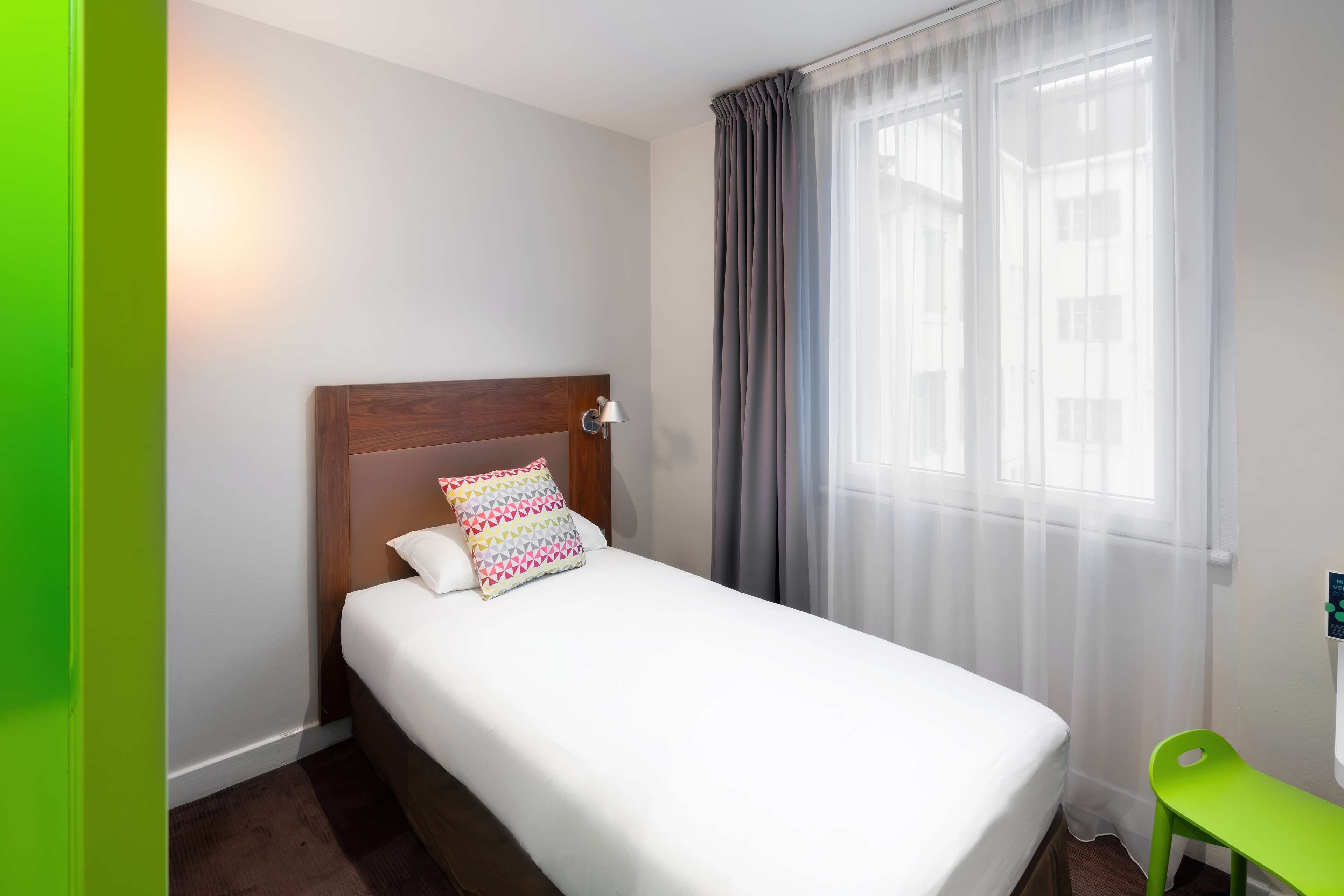 A hotel room with a single bed, a bedside lamp, a green chair, and a window with sheer curtains.