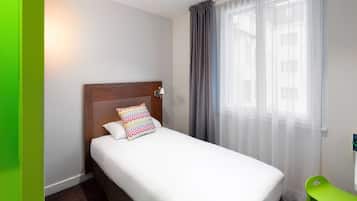 A hotel room with a single bed, a bedside lamp, a green chair, and a window with sheer curtains.