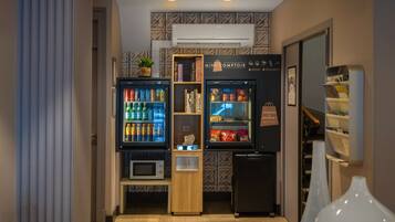 Una kitchenette compacta con dos mini refrigeradores, un microondas y una pequeña estantería con libros y una planta.
