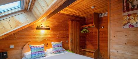 A cozy wooden cabin bedroom with a sloped ceiling, a bed with colorful pillows, a small wooden bench, and a wall-mounted coat rack.