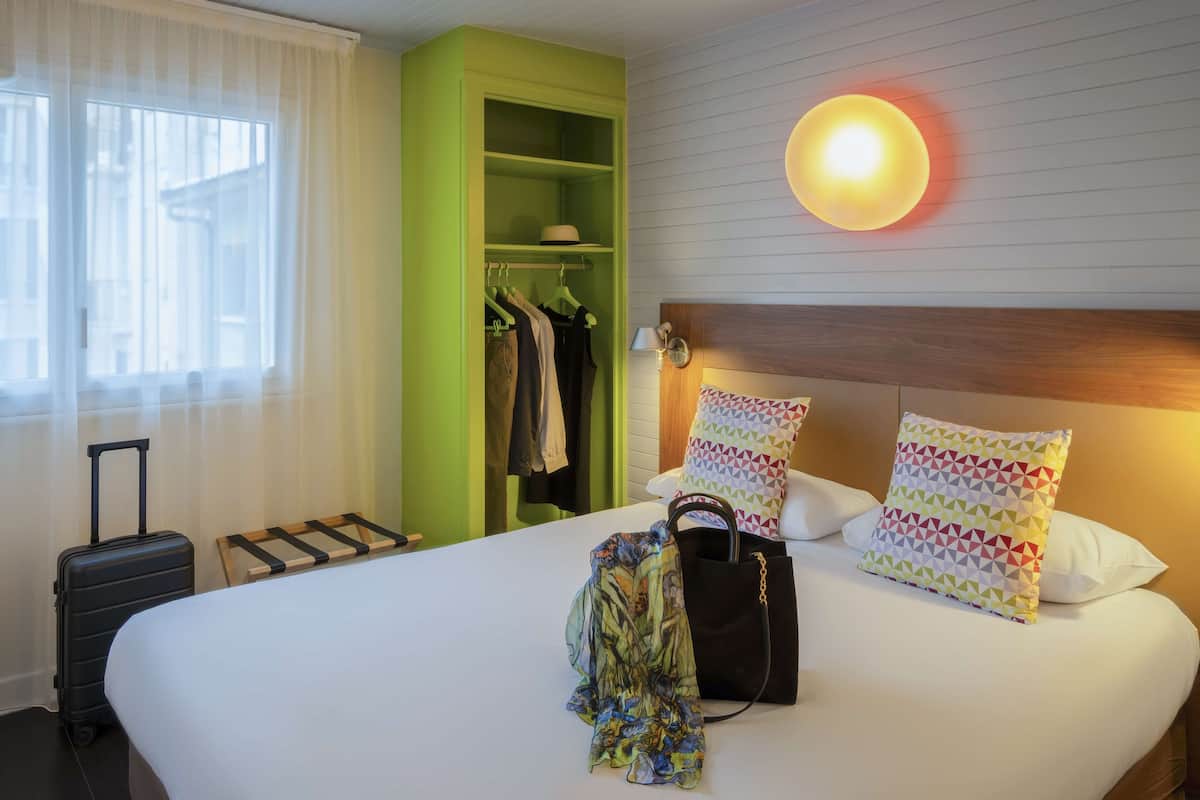 Una habitación de hotel con cama, armario, maleta y perchero.