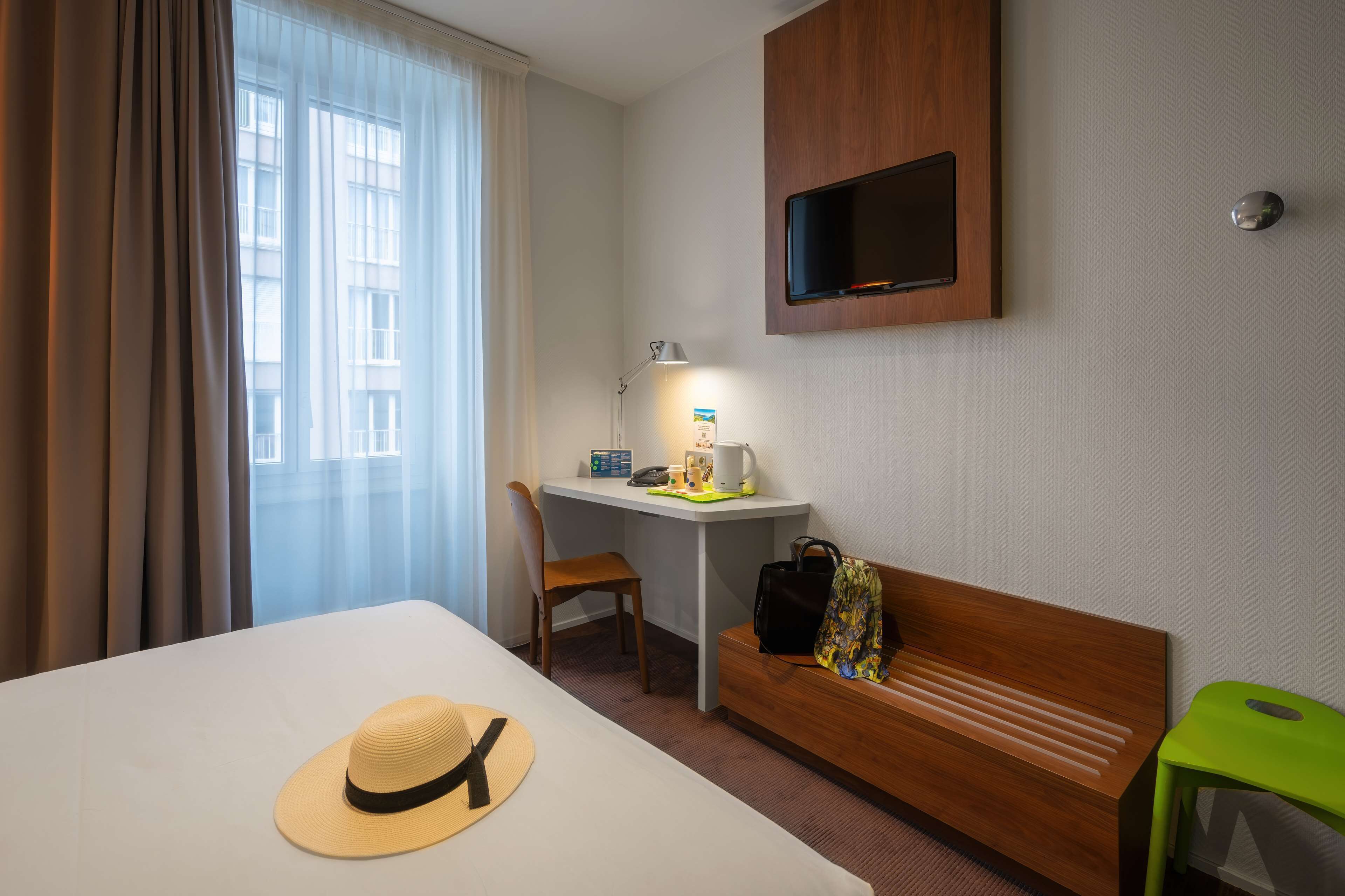 Quarto de hotel com uma cama, uma escrivaninha, uma cadeira, uma TV e um banquinho verde.