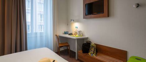 A hotel room with a bed, a desk, a chair, a TV, and a green stool.