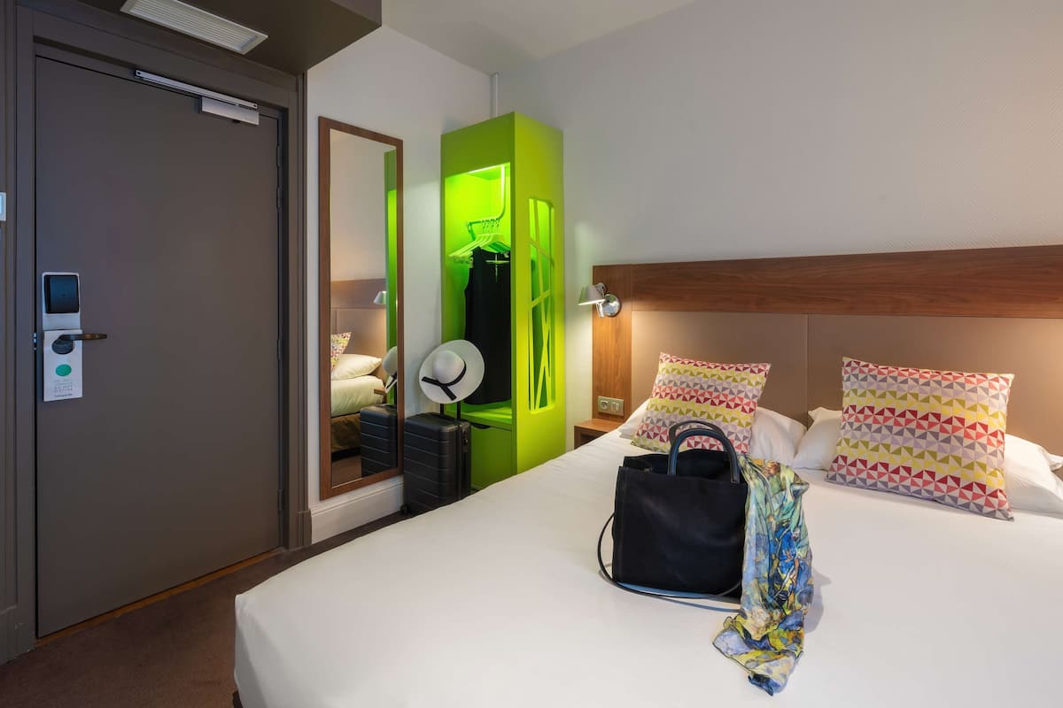 Una habitación de hotel con una cama, un armario verde, un espejo, una mesita de noche y una lámpara de pared.
