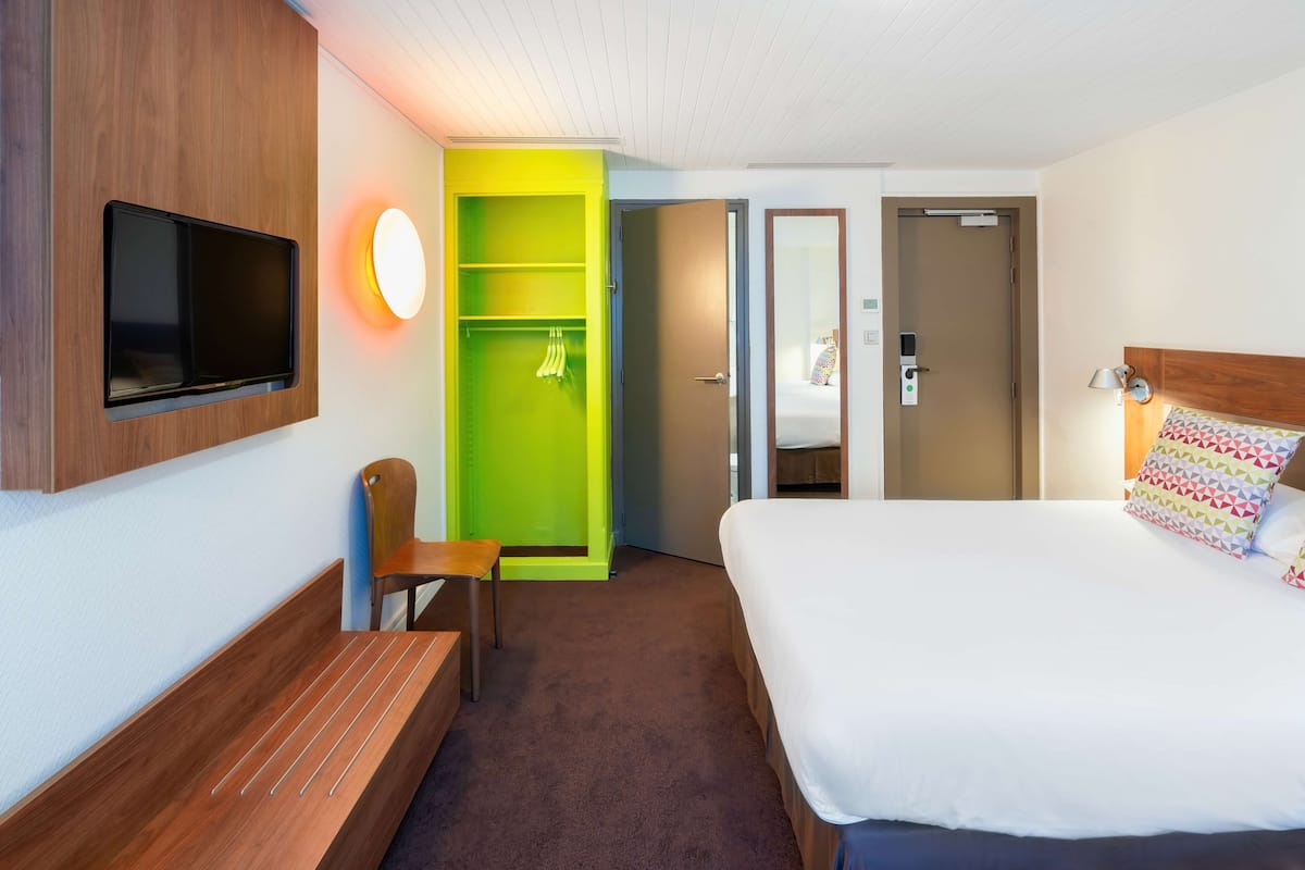 Habitación de hotel con cama, televisor montado en la pared, zona de estar con un banco y una silla, un mueble verde de almacenamiento y una puerta que da a otra habitación.