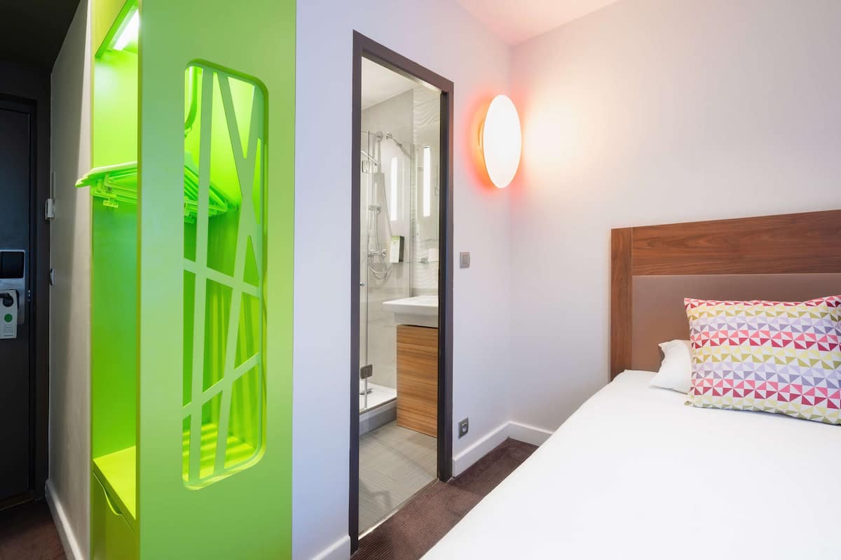 Habitación de hotel con cama, mesita de noche, espejo y baño visible a través de una puerta abierta.