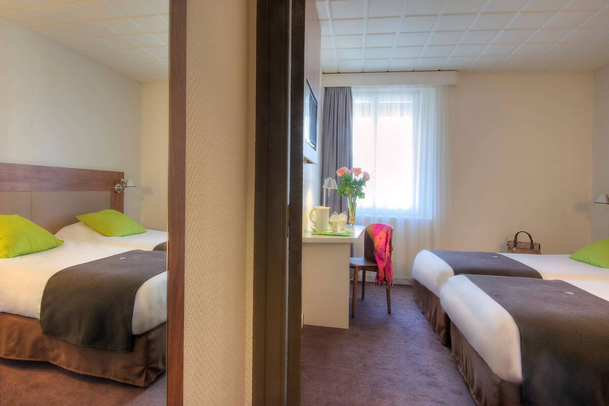 Una habitación de hotel con dos camas, un escritorio con silla, una ventana con cortinas y un jarrón con flores.