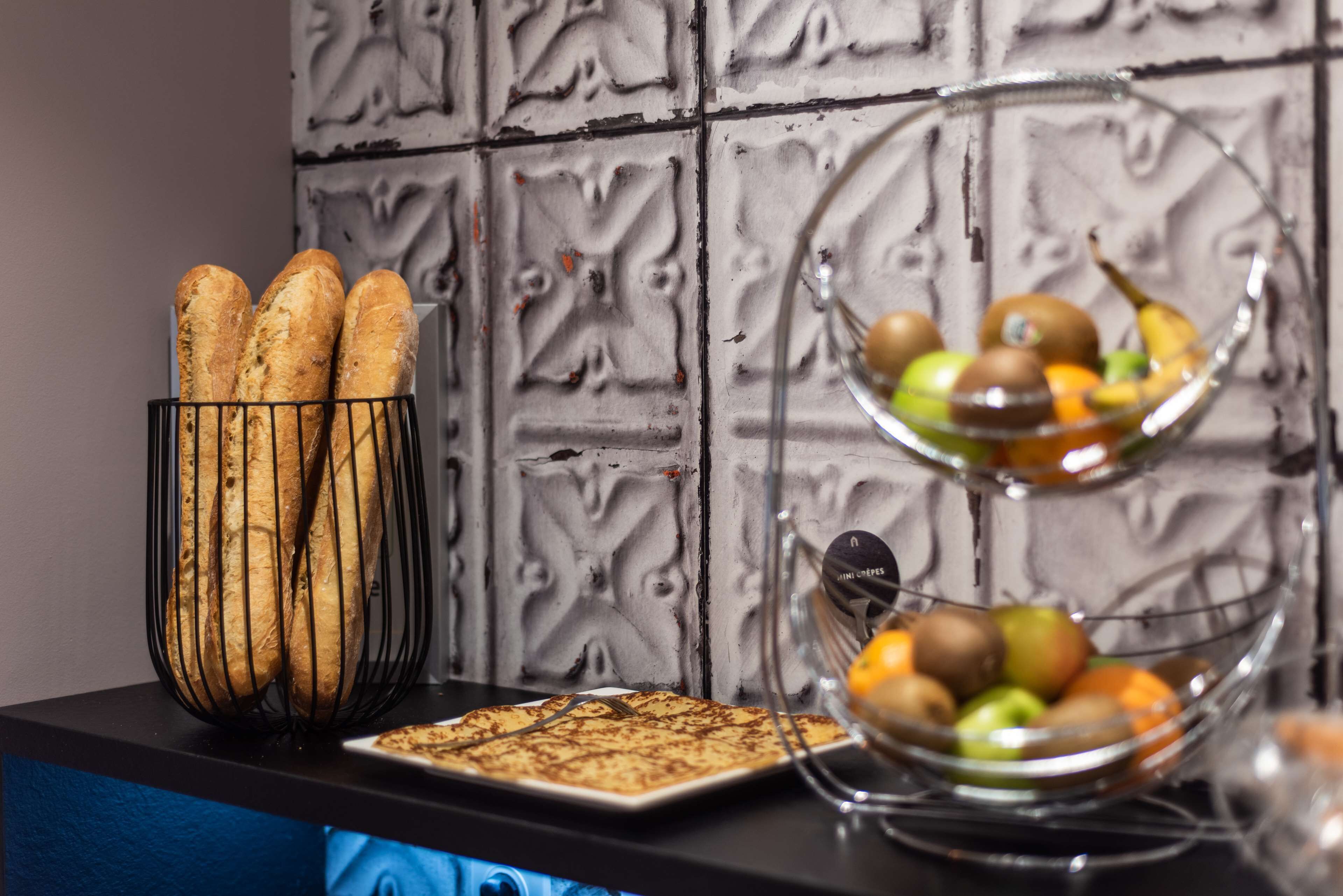 Een keukenblad met stokbroden in een metalen mand, een bord met een gebakje en een fruitschaal met appels en sinaasappels.