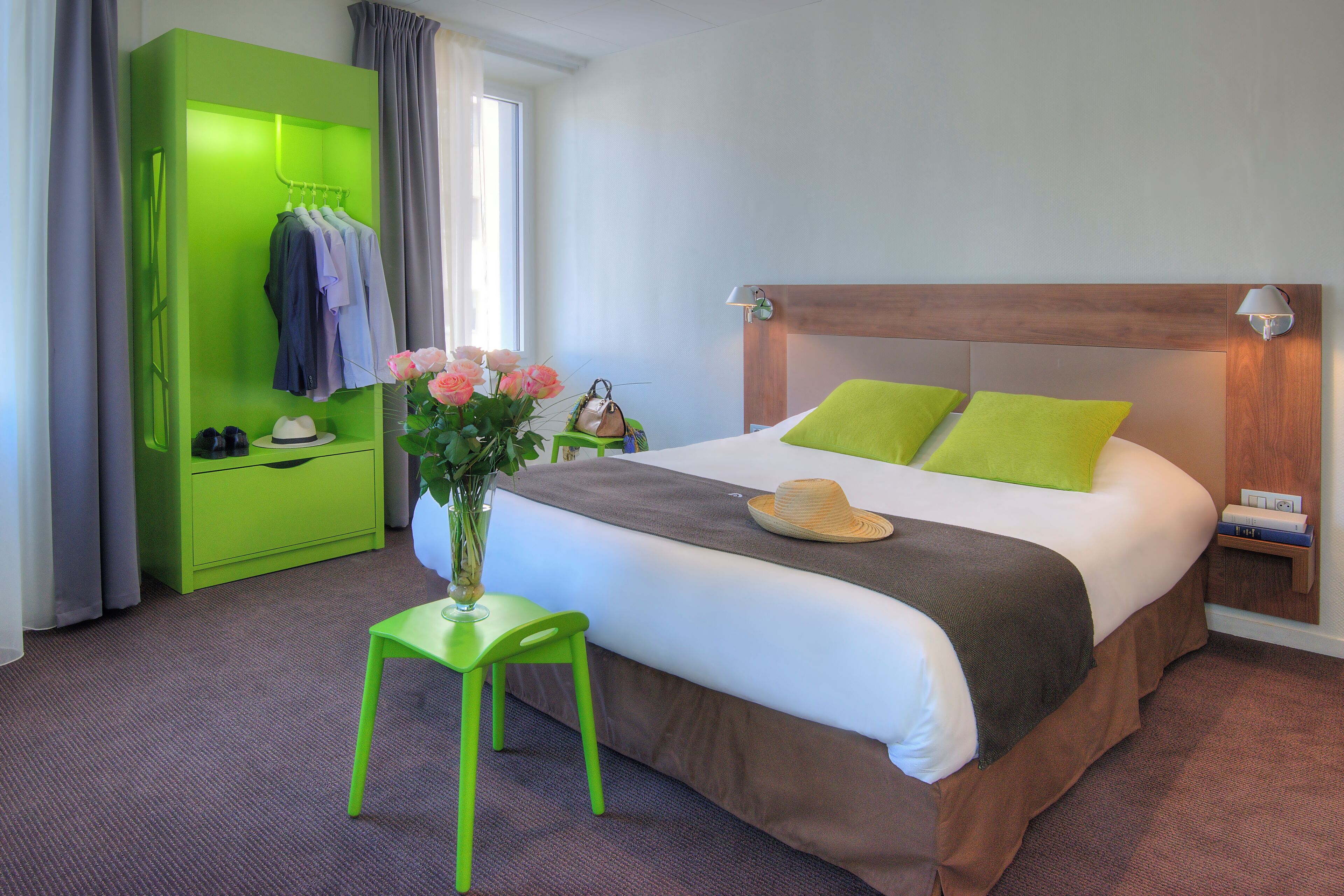 A hotel room with a bed, a green wardrobe, a nightstand, a chair, and a vase with flowers.