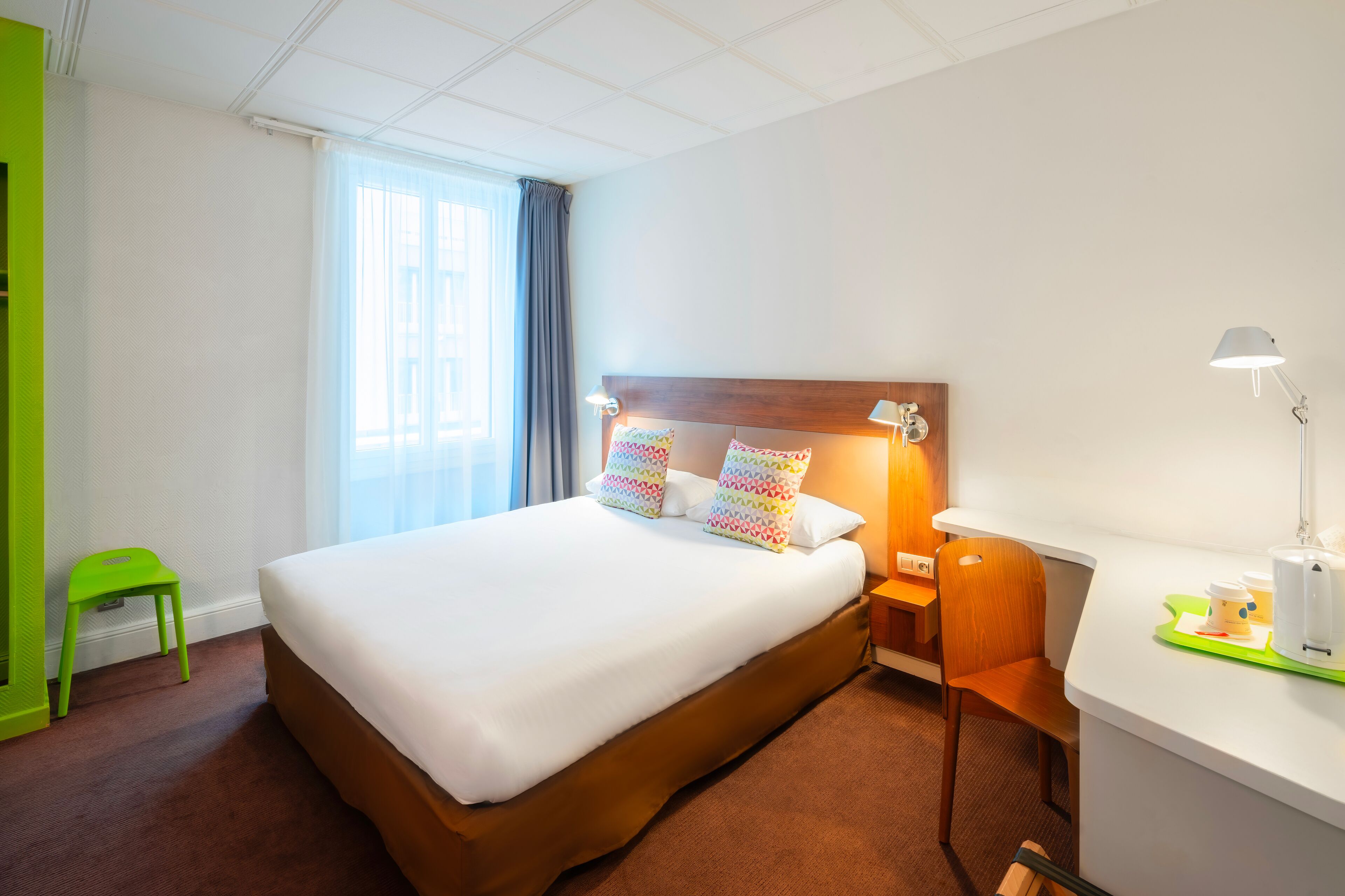Une chambre d’hôtel avec un grand lit, un bureau avec une lampe, une chaise, un petit tabouret et une fenêtre avec des rideaux.