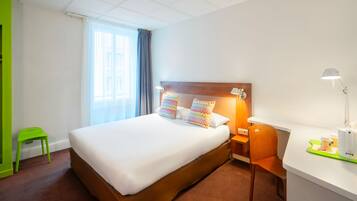 Une chambre d’hôtel avec un grand lit, un bureau avec une lampe, une chaise, un petit tabouret et une fenêtre avec des rideaux.