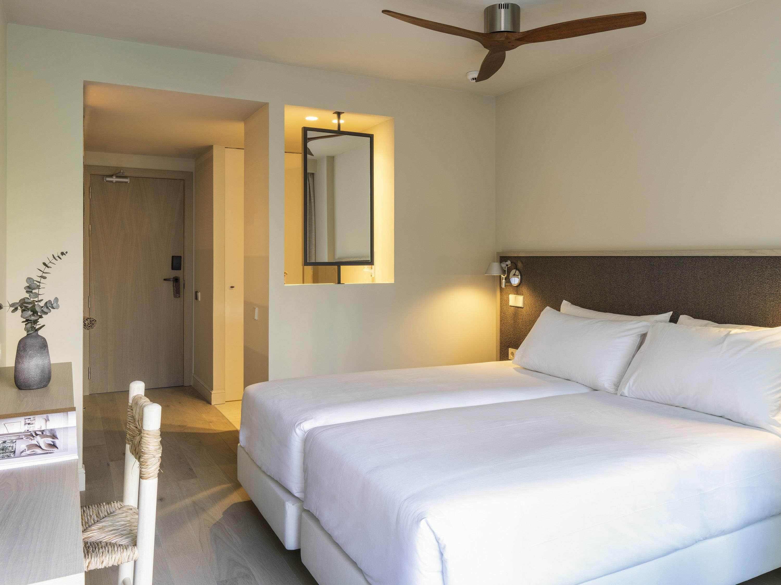 A hotel room with a bed, a desk, a chair, a mirror, and a ceiling fan.
