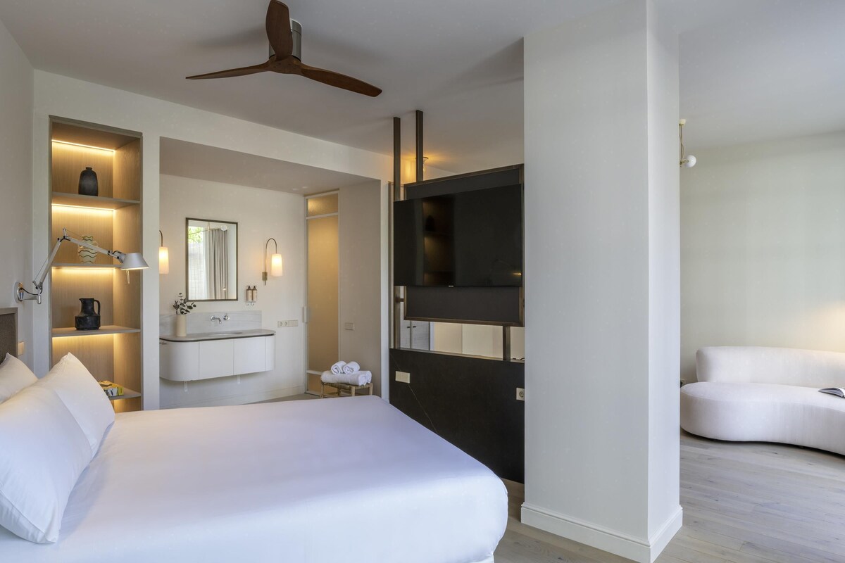 A modern hotel room with a bed, a vanity with a mirror, a ceiling fan, and a seating area.