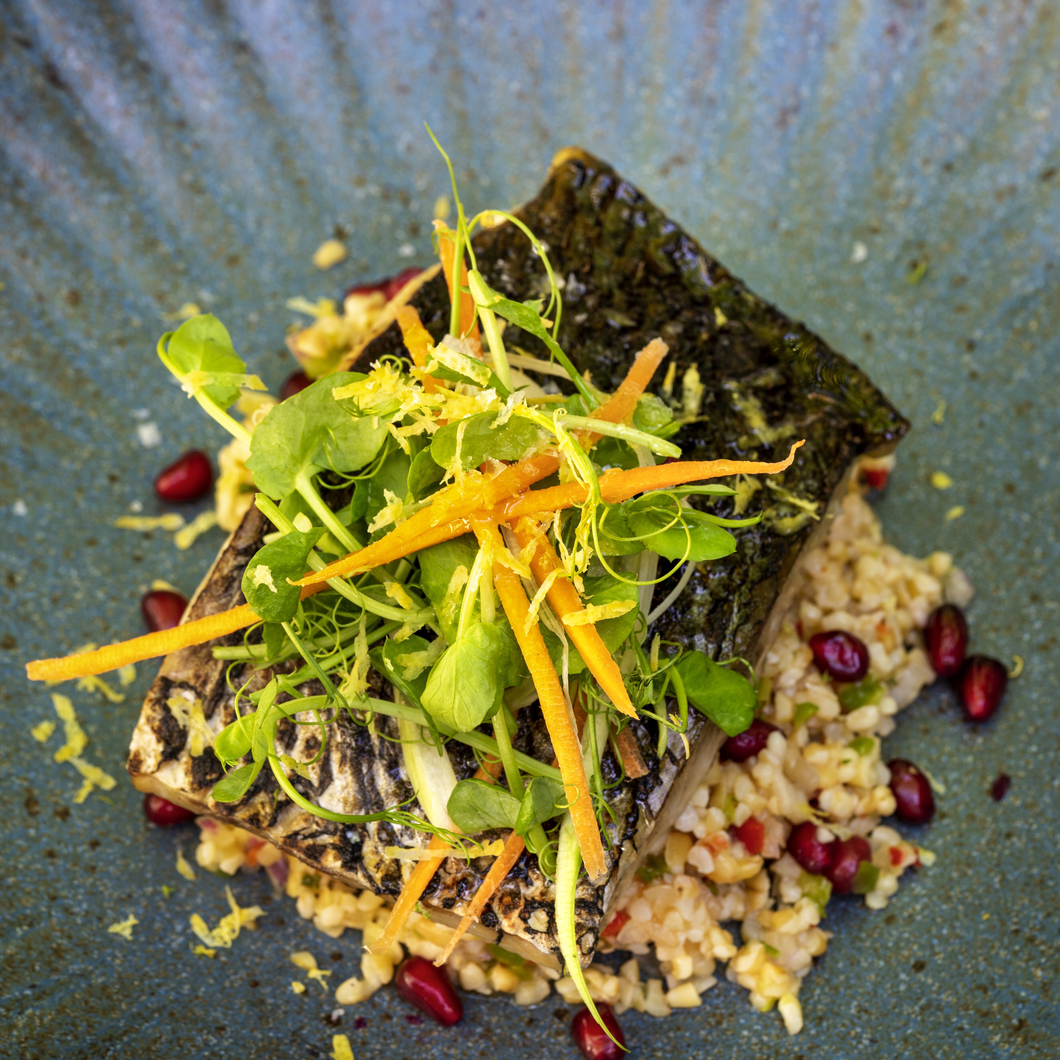 Un plato con un trozo de pescado cubierto de vegetales y zanahoria rallada, servido sobre un plato azul con textura y semillas de granada esparcidas.