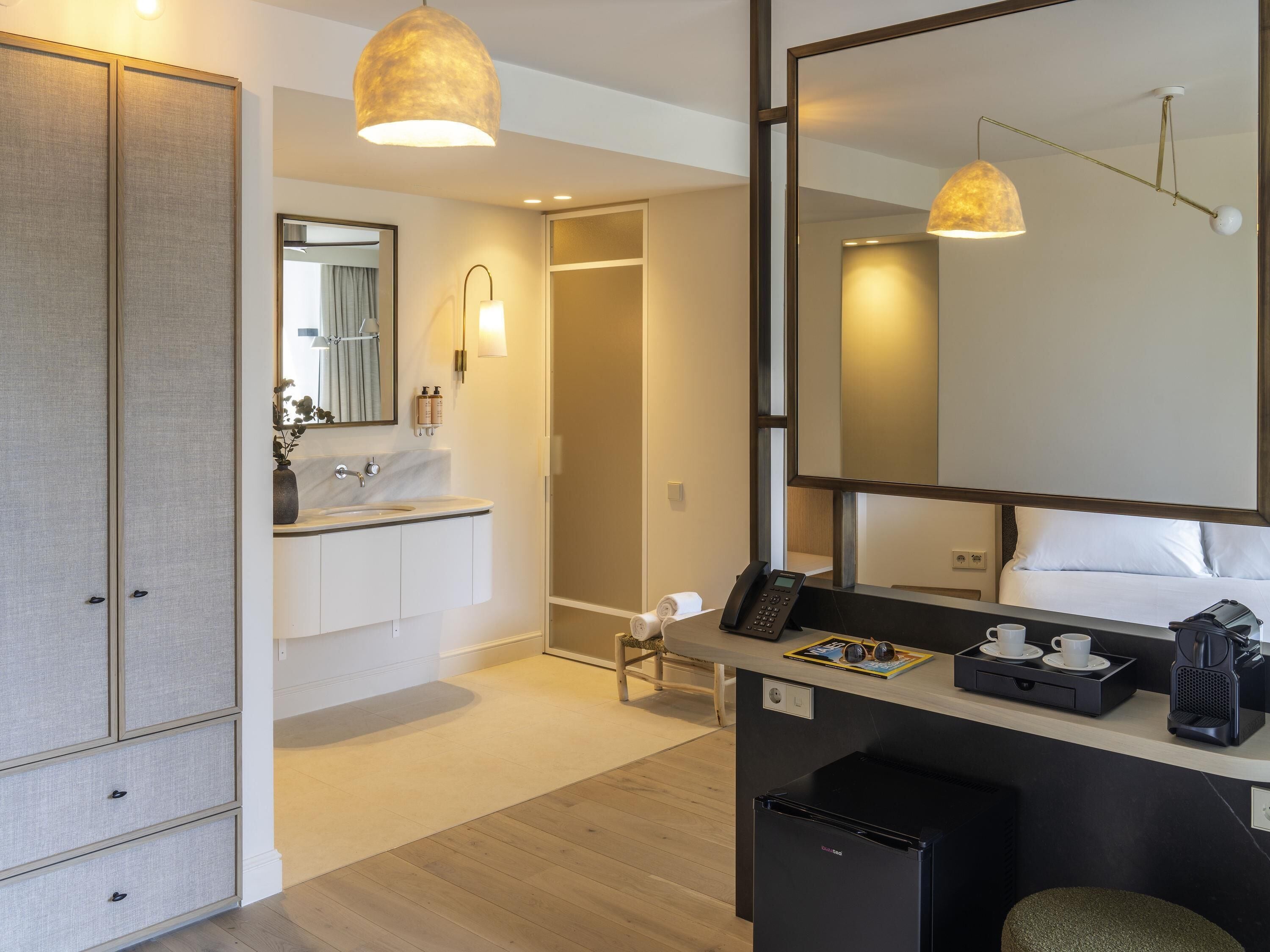 A modern hotel bathroom with a large mirror, a sink, a toilet, and a small seating area with a coffee machine.