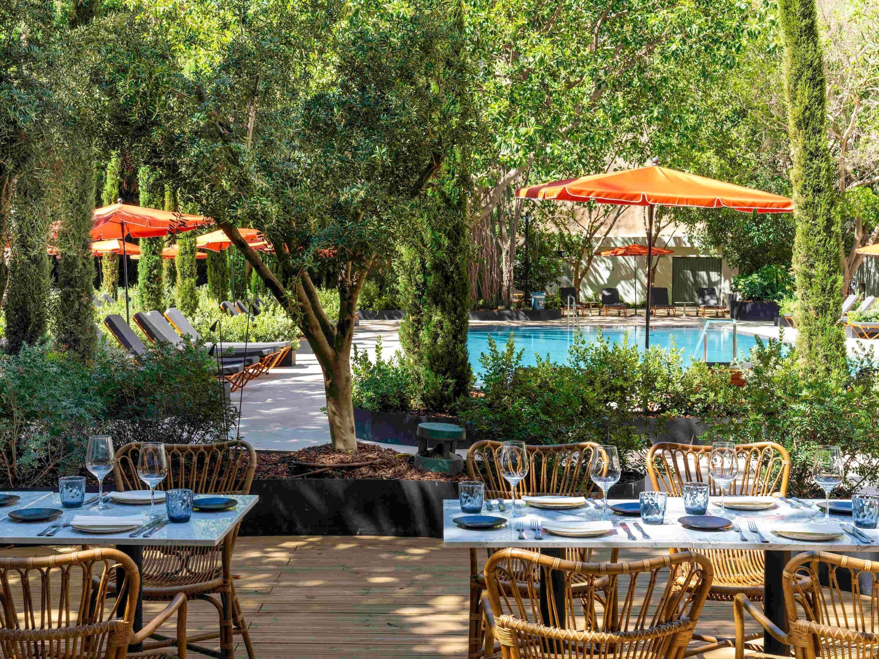Un espace extérieur aménagé pour les repas, avec des chaises et des tables en osier, une piscine et des parasols rouges.