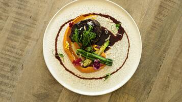 A round plate with a dark center, surrounded by a light-colored sauce and garnished with green vegetables and flowers.