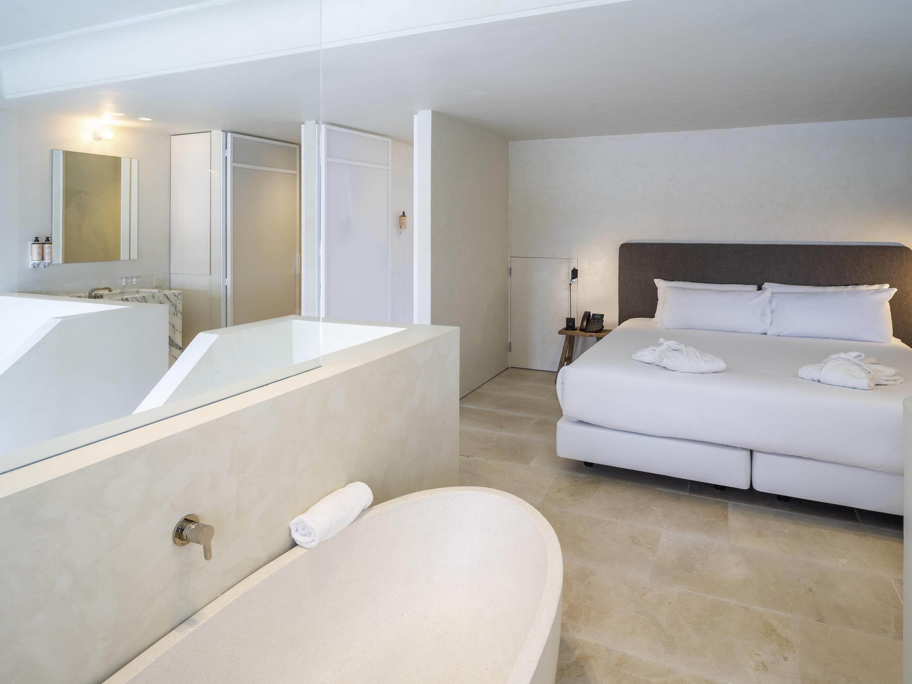 Ein modernes Hotelzimmer mit einer großen Badewanne, einer separaten Duschkabine, einem Bett mit weißer Bettwäsche und einem Nachttisch mit Lampe und Telefon.