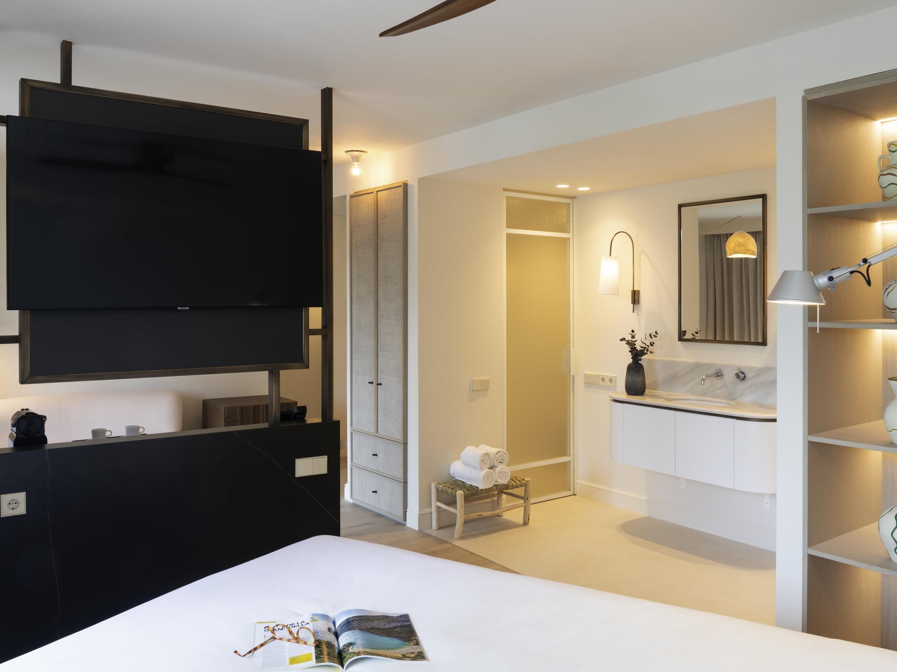 A modern hotel room with a bed, a TV, a bathroom with a sink and mirror, and a small table with towels.