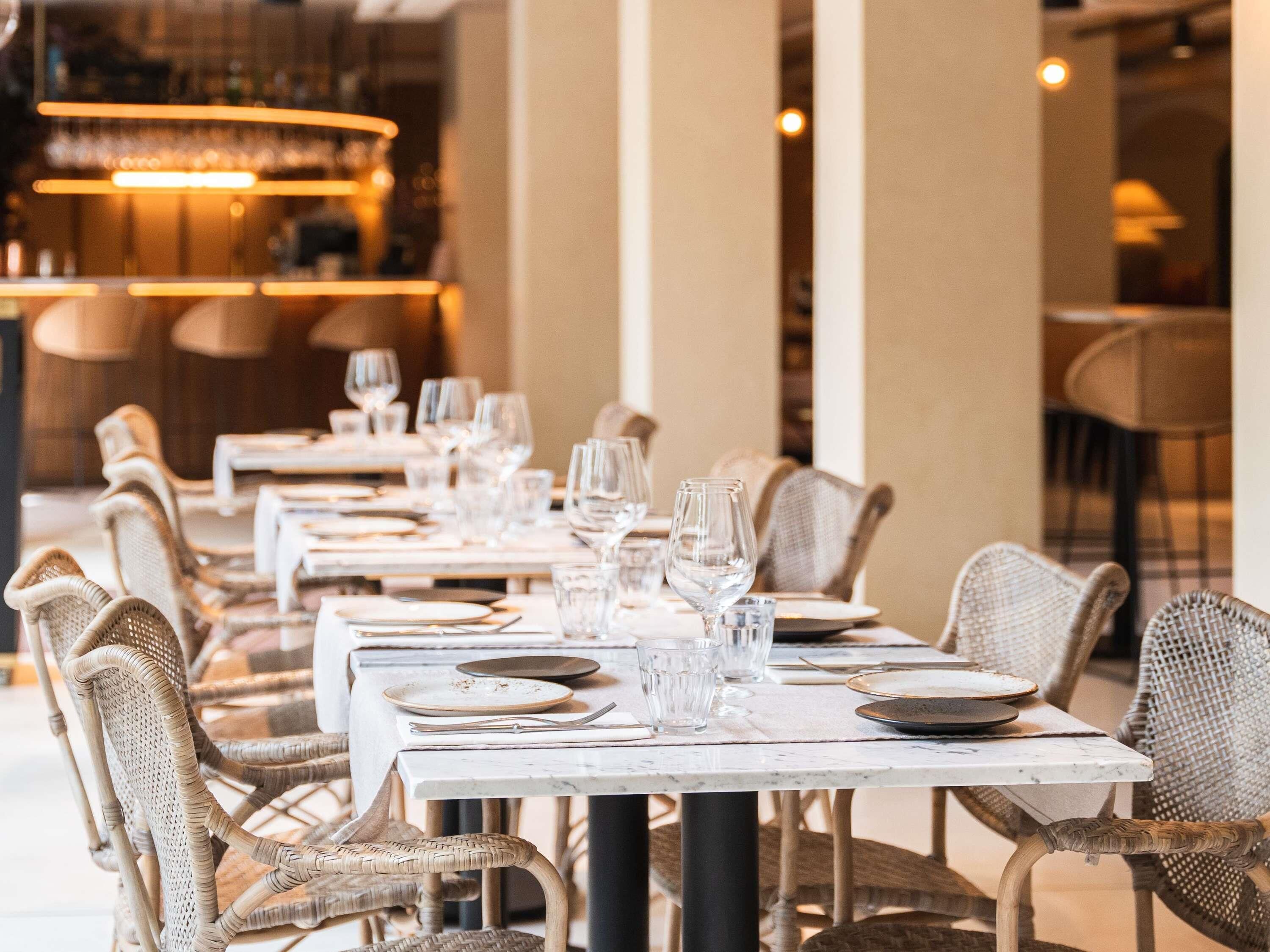 A restaurant with multiple tables set for dining, featuring wicker chairs and a modern chandelier.
