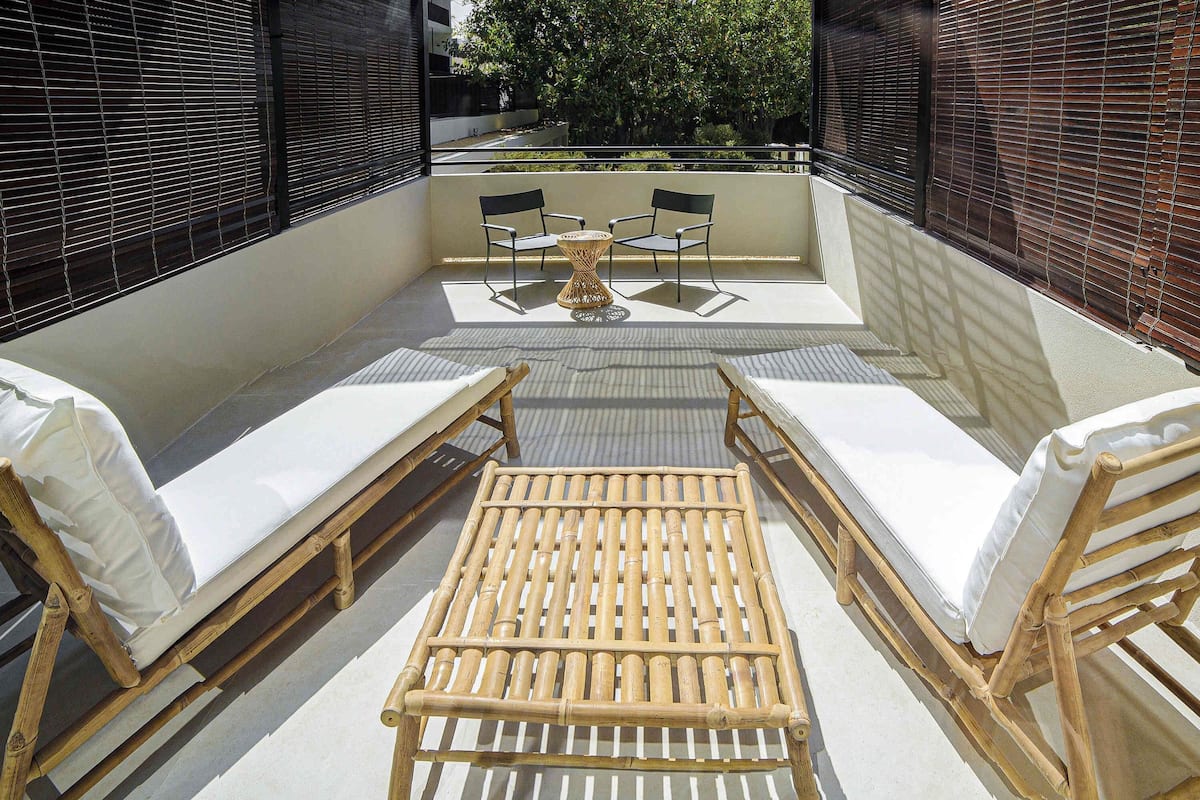 A sunlit outdoor seating area with a wooden table and chairs, a small round table, and a view of the street and greenery.