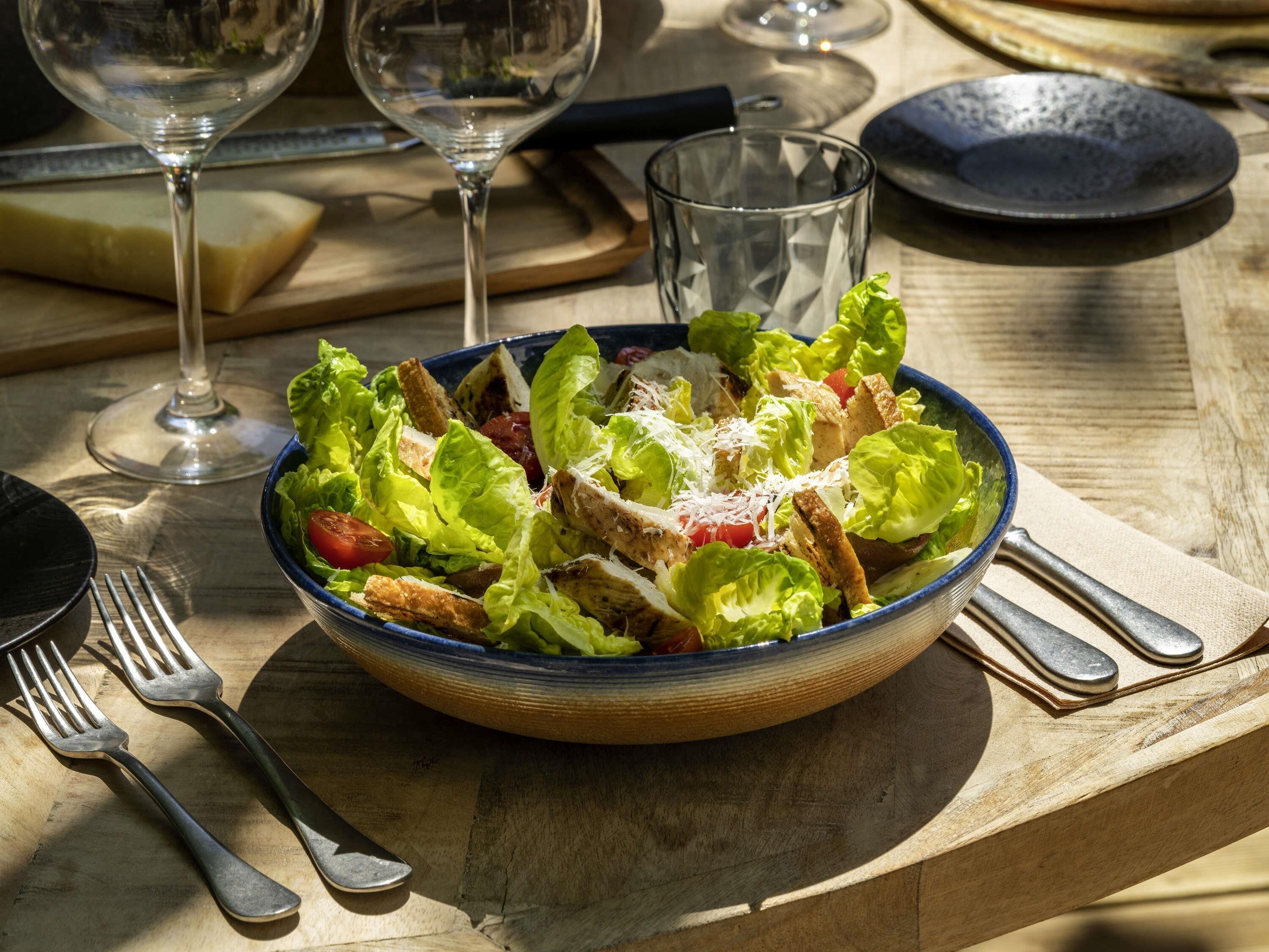 Un cuenco de ensalada sobre una mesa de madera, con tenedores y una copa de vino.