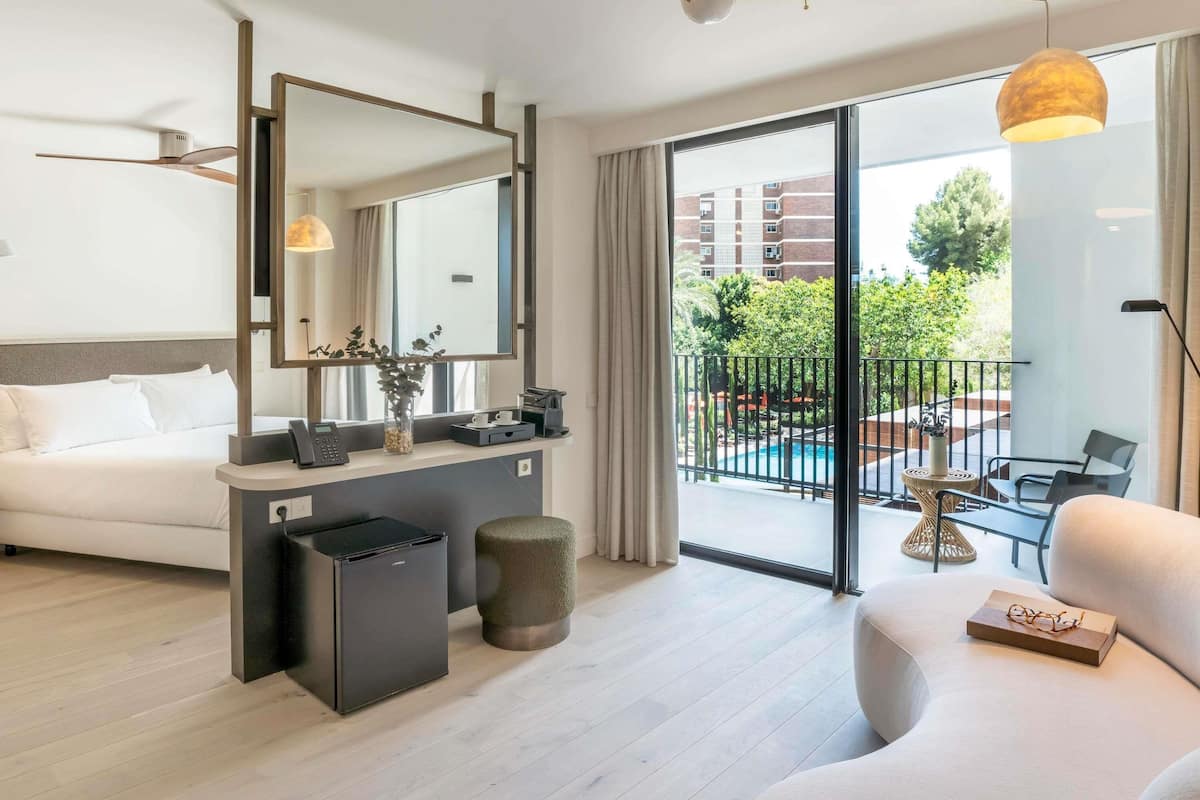 A hotel room with a bed, a desk, a chair, a small trash bin, and a balcony with a pool.