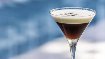Un bicchiere da martini con un liquido scuro e una schiuma, appoggiato su una superficie bianca su sfondo blu sfocato.