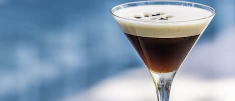 Ein Martini-Glas mit dunkler Flüssigkeit und Schaumkrone auf einer weißen Oberfläche vor unscharf blauem Hintergrund.