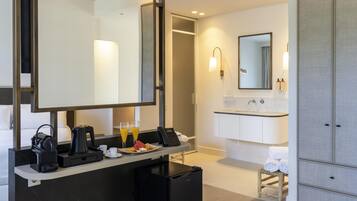 A modern hotel bathroom with a large mirror, a sink, and a small refrigerator.
