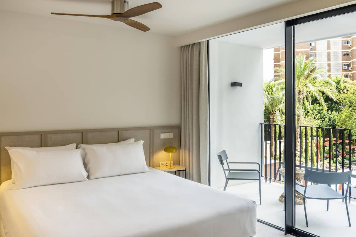 A hotel room with a bed, a nightstand, a lamp, a ceiling fan, and a balcony with outdoor furniture.