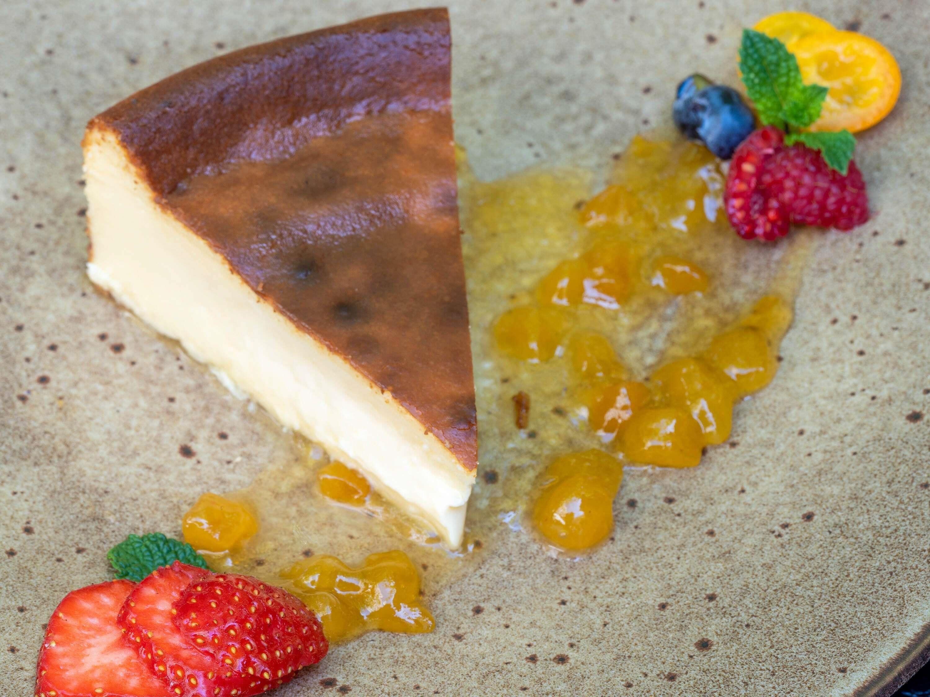 Une part de gâteau au fromage, avec une croûte dorée, servie avec un assortiment de fruits rouges frais et un filet de sauce à l’orange.