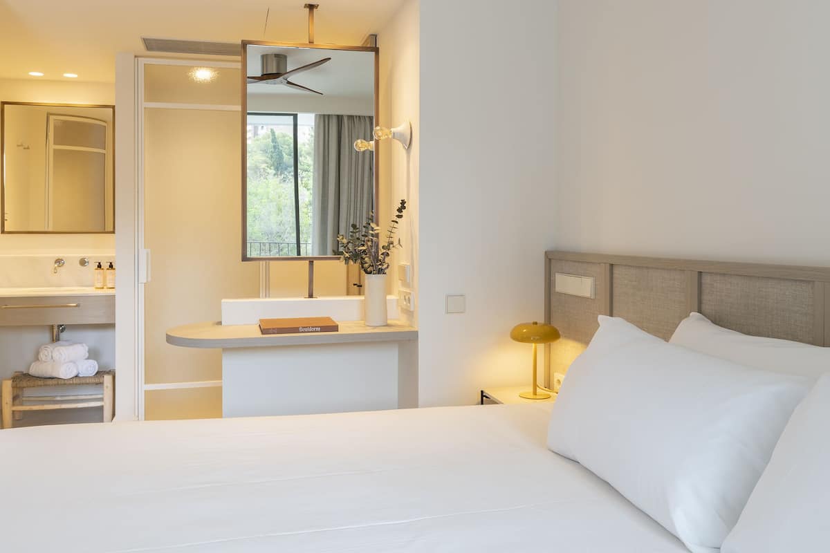 A hotel room with a bed, a bedside table with a lamp, a vanity with a mirror, a towel rack, and a window with curtains.