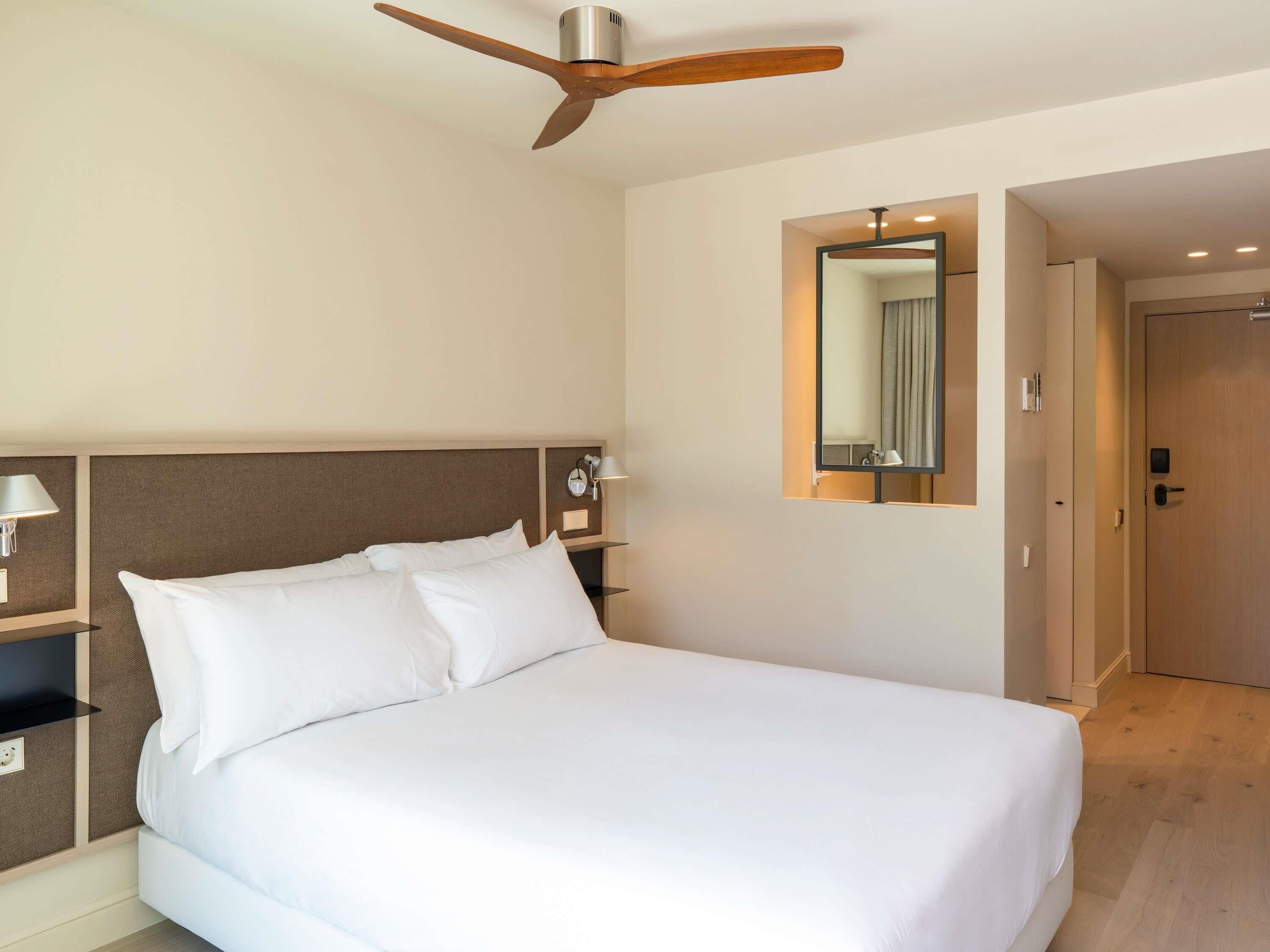 A hotel room with a bed, a ceiling fan, a nightstand with a lamp, a mirror, and a door.