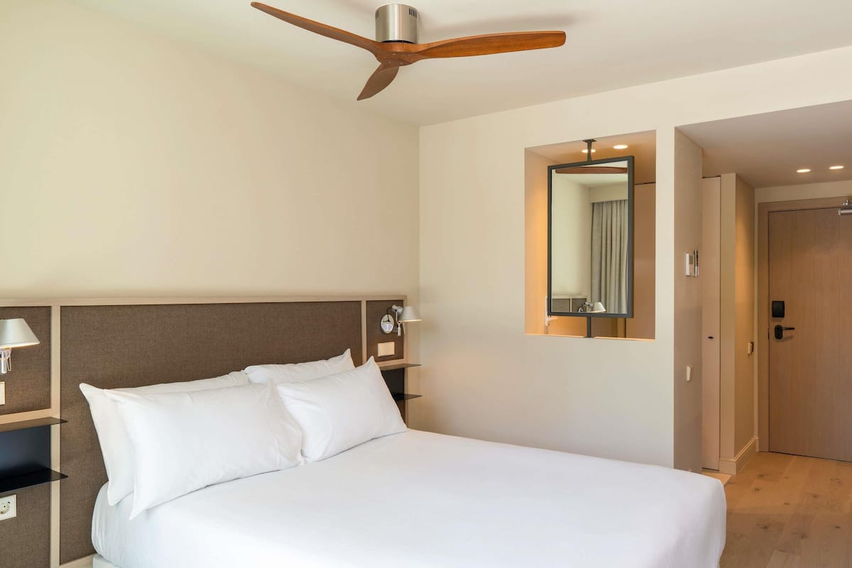 A hotel room with a bed, a ceiling fan, a nightstand with a lamp, a mirror, and a door.