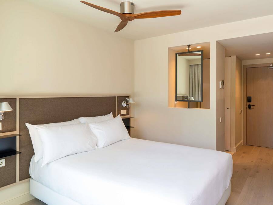 Una habitación de hotel con una cama, ventilador de techo, mesita de noche con lámpara, espejo y una puerta.
