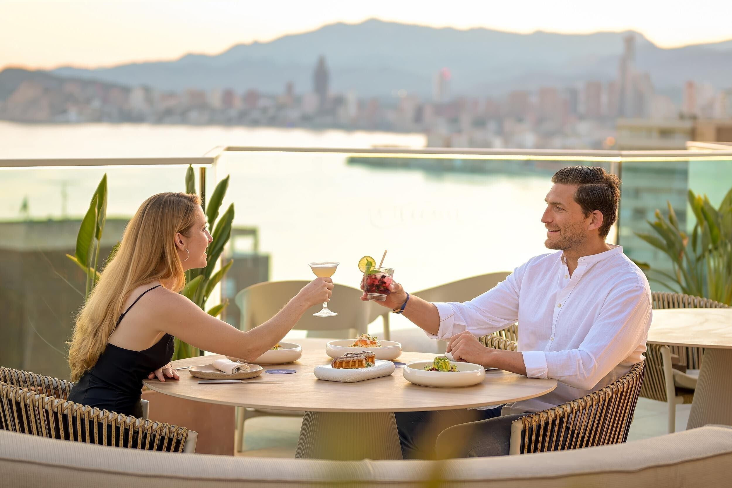 Una pareja cenando en una mesa al aire libre con vista a la ciudad, rodeada de mobiliario de mimbre y una mesa con platos, vasos y un cóctel.