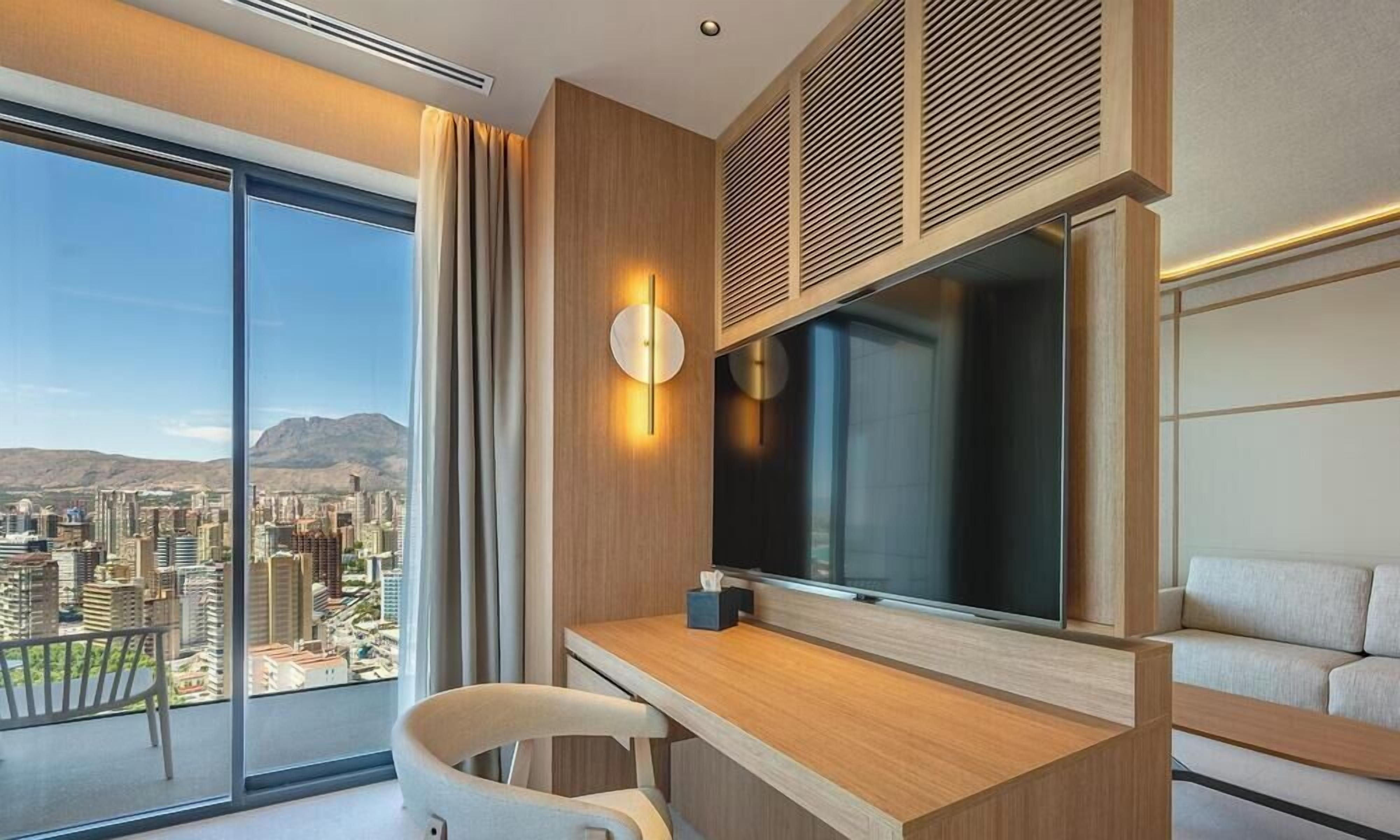 Una habitación de hotel moderna con un ventanal grande, un televisor de pantalla plana, un escritorio y un sofá.